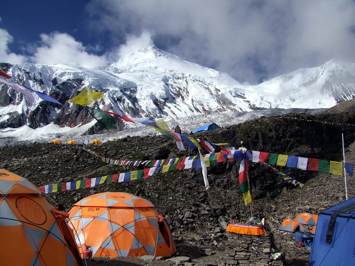 A vibrant orange geodesic dome tent sits at a high altitude base camp with colorful prayer flags and snowy peaks during a Luxury Manaslu Trek.