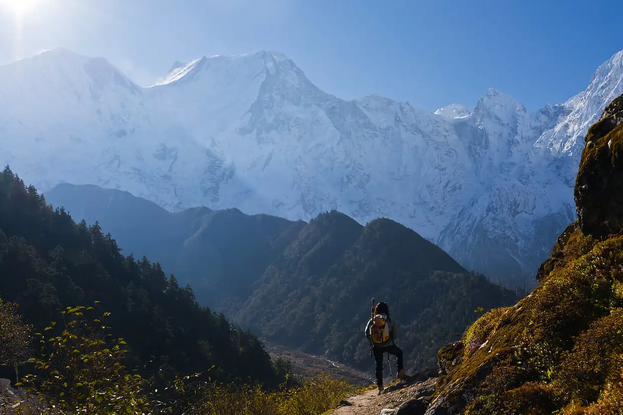 A lone trekker stands on a mountain path looking at massive snow covered peaks under a bright sun during a Luxury Manaslu Trek.