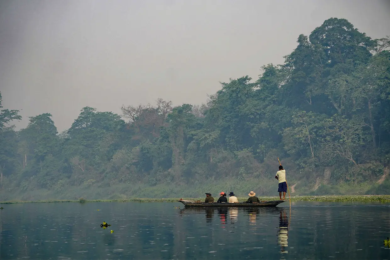 Travelers enjoy a peaceful river safari in a traditional dugout canoe during a Luxury Kathmandu Pokhara Chitwan Tour. A local guide poles through calm waters by a misty forest.