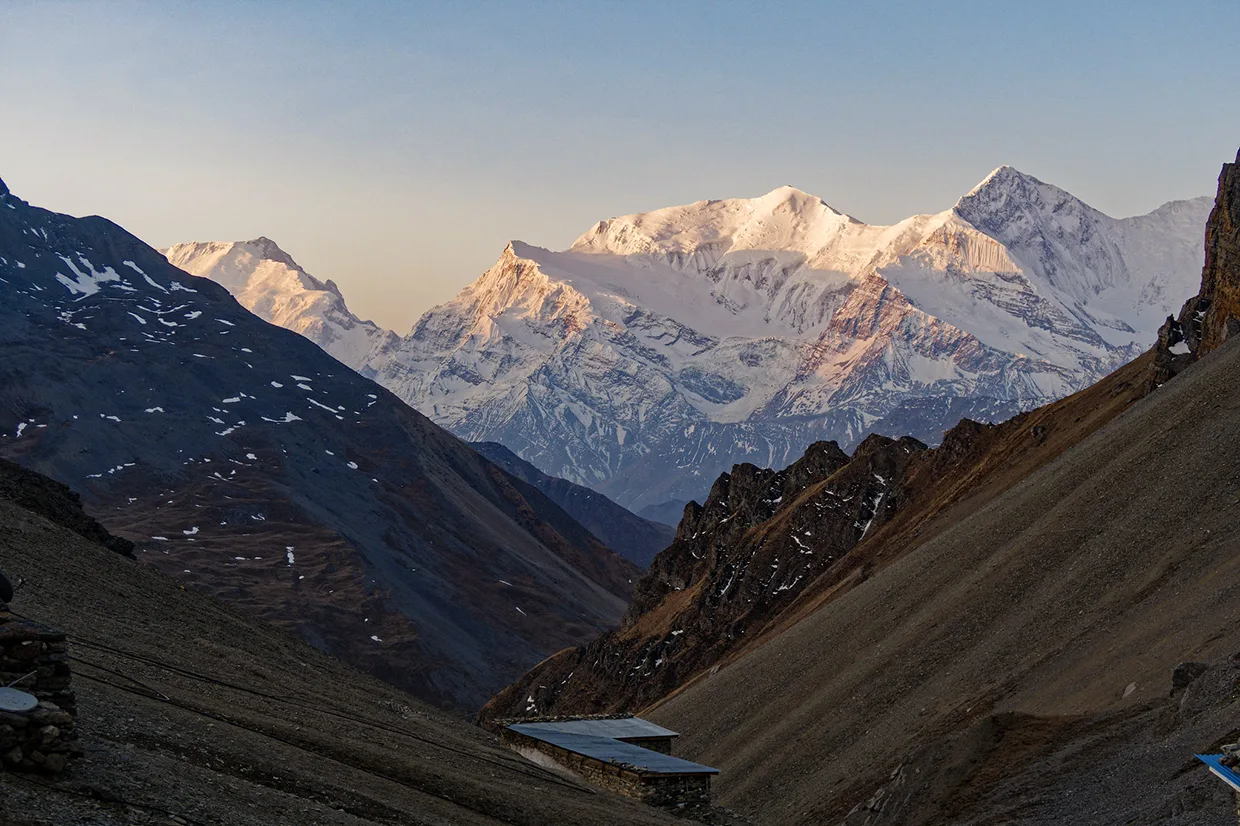 Golden sunlight illuminates snow-dusted Himalayan peaks above a high mountain lodge nestled in a deep valley on a Luxury Annapurna Circuit Trek.