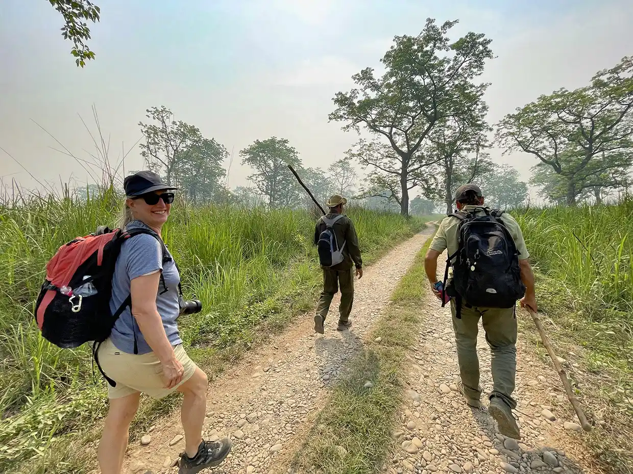 A smiling woman walks on a sunny trail with two guides during a Luxury Kathmandu Pokhara Chitwan Tour. Tall grass and trees line the dirt path.