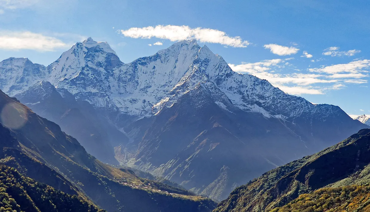 A wide view of snow-dusted Himalayan peaks rising above a lush valley with a small mountain village nestled below on the Luxury Everest Base Camp & Gokyo Lakes Trek.