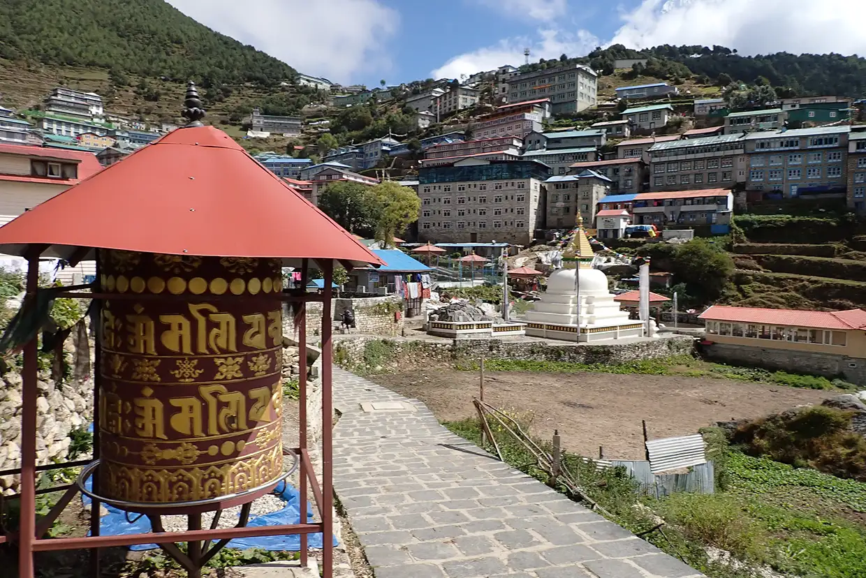 A large decorative prayer wheel stands in the foreground of Namche Bazaar, with a white stupa and mountain lodges visible during a Luxury Everest Base Camp & Gokyo Lakes Trek.