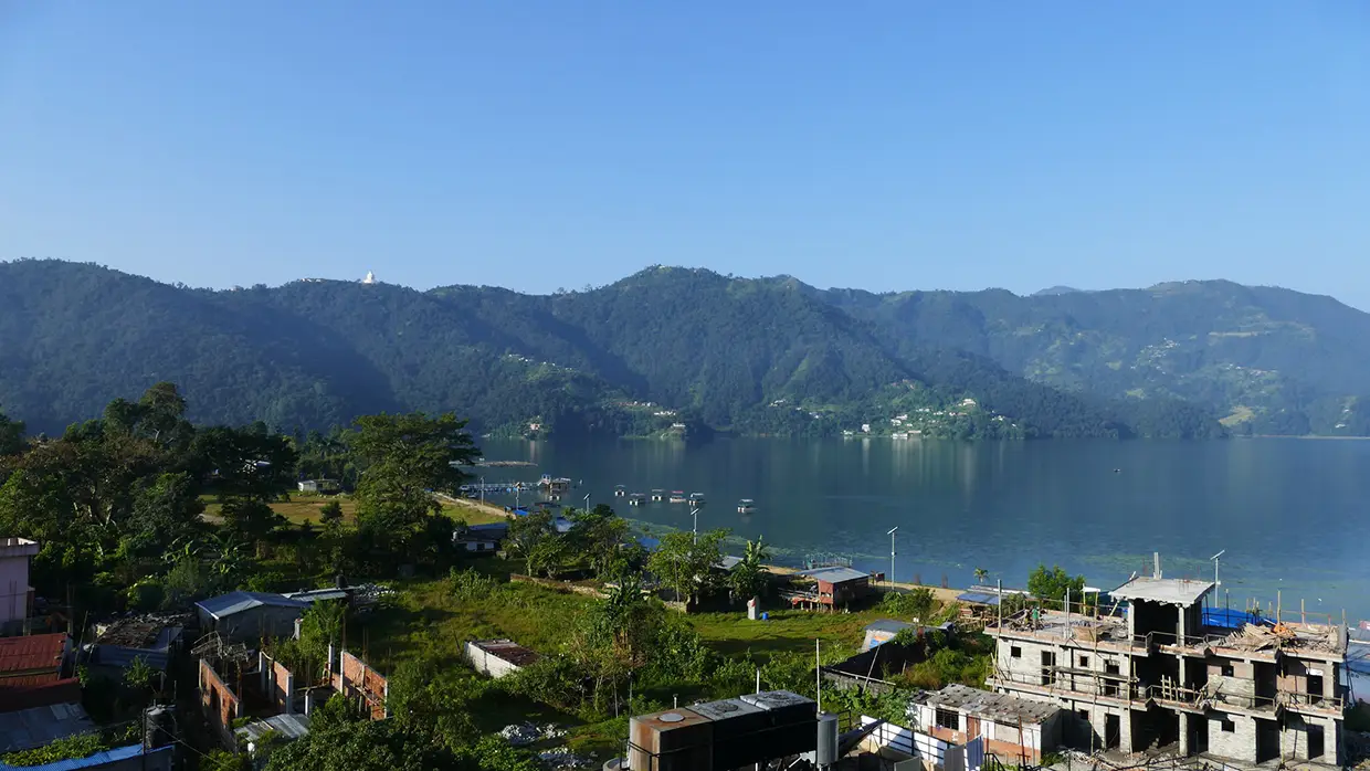 A calm lake surrounded by green mountains and village buildings provides a peaceful rest stop during your Kathmandu Luxury Experience in the Himalayas.