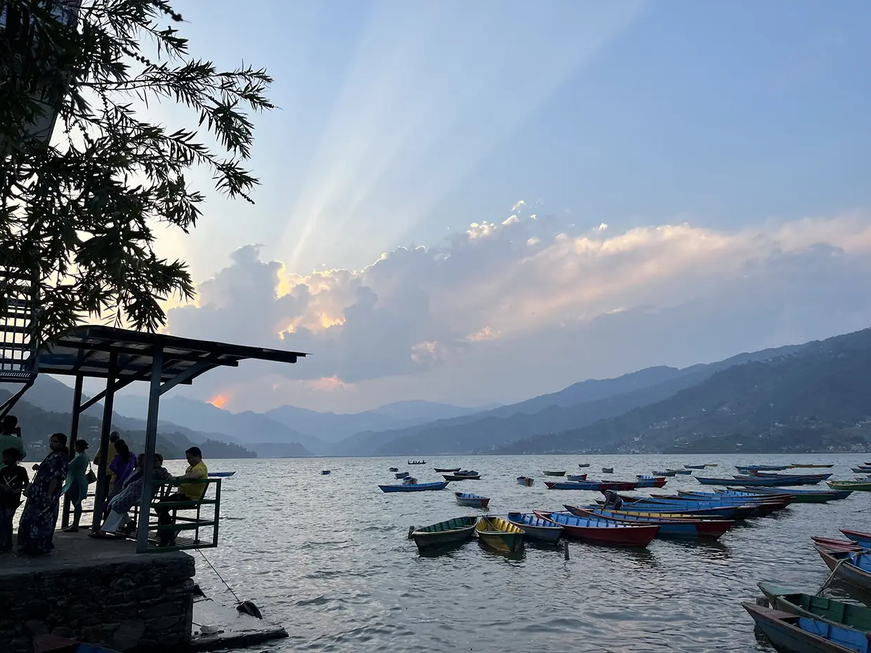 Boats rest on the shore of a lake at sunset while people relax on a dock during a Luxury Kathmandu Pokhara Chitwan Tour.