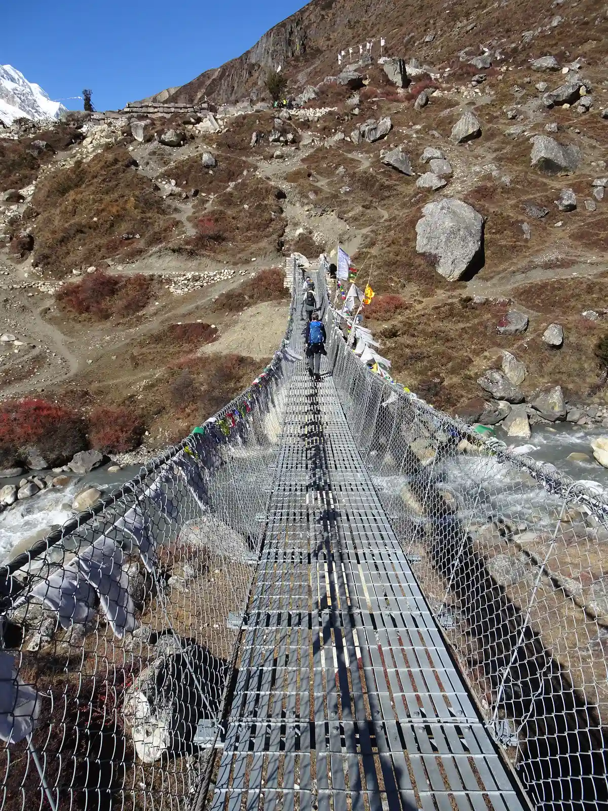 Trekkers cross a long metal suspension bridge draped with prayer flags over a mountain river during a Luxury Manaslu Trek in Nepal.