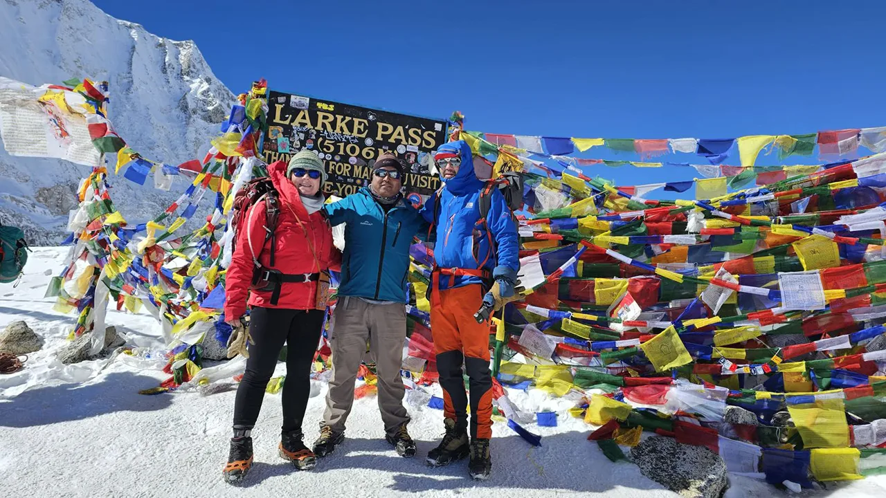 Guide and trekkers posing at Larke La Pass surrounded by colorful prayer flags and snow-covered Himalayan peaks.