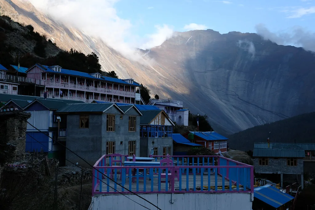 Traditional mountain lodges and colorful balconies in the village of Upper Pisang sit beneath towering, sun-lit cliffs on a Luxury Annapurna Circuit Trek.