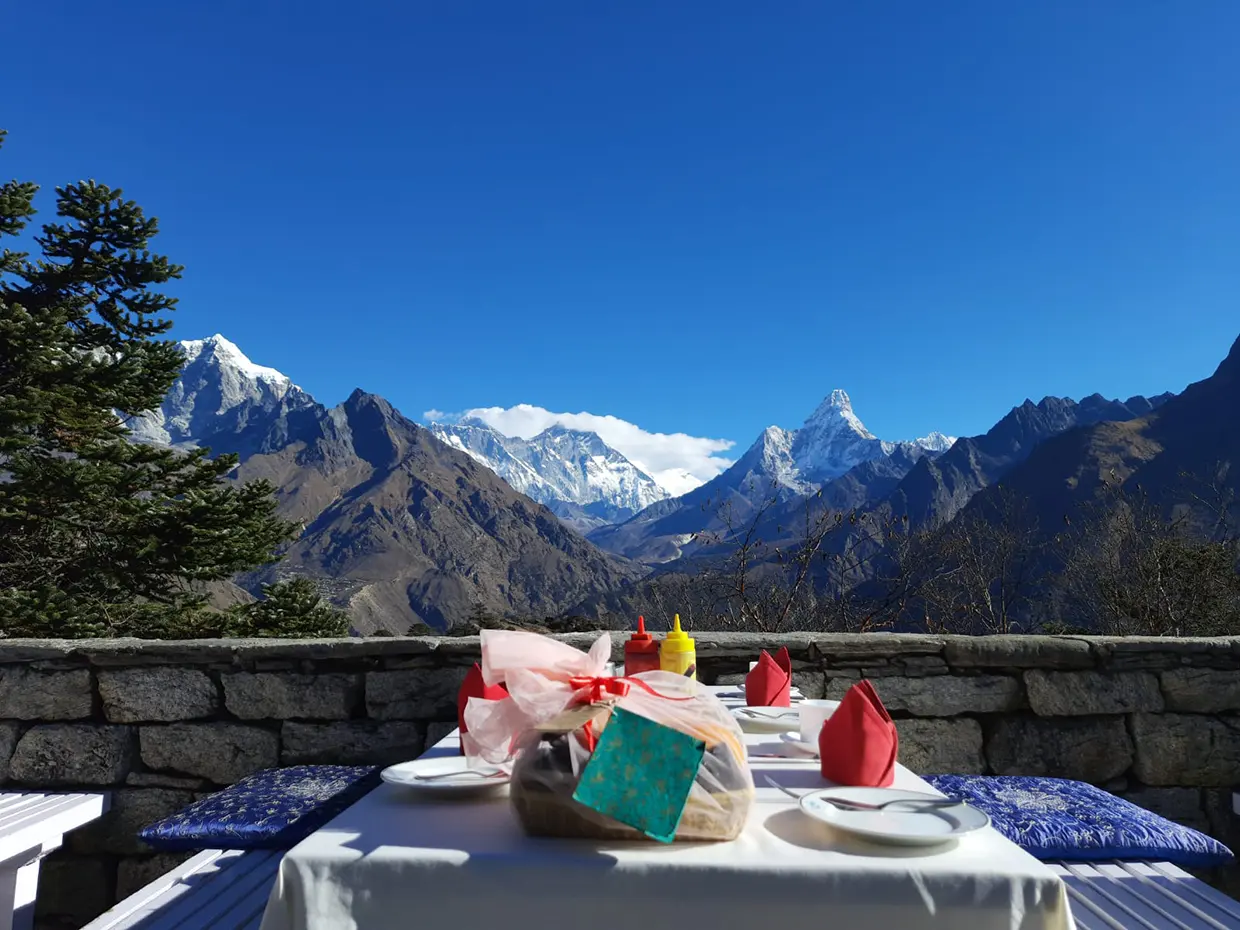 A luxurious outdoor dining table set with white linen, red napkins, and blue cushions, overlooking stunning snowy Himalayan peaks under a clear sky, a curated stop on our Luxury Nepal Honeymoon Tour.