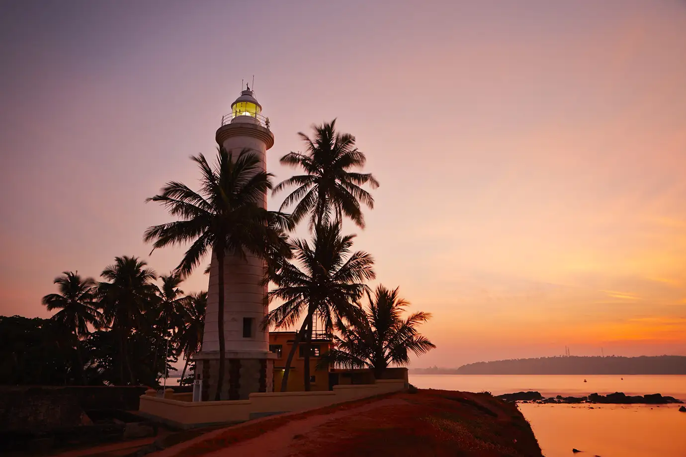 A tall white lighthouse with an illuminated beacon stands among silhouetted palm trees against a backdrop of a vibrant pink, purple, and orange sunset sky. The lighthouse sits on a coastal path next to calm ocean waters with a rocky shoreline.