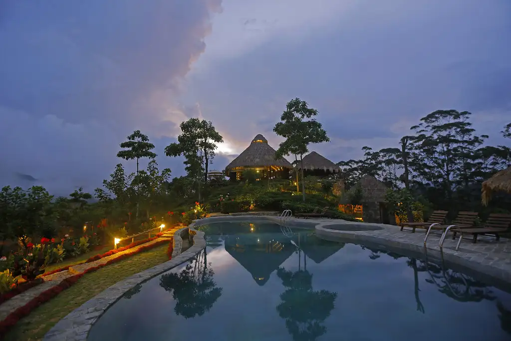 A tranquil swimming pool at dusk reflecting a luxury resort with traditional thatched-roof villas perched on a lush, terraced hill. Warm yellow lights illuminate the garden pathways and the interiors of the pavilions. The sky is a moody blue and purple, with a bright, glowing break in the clouds directly behind the central villa.