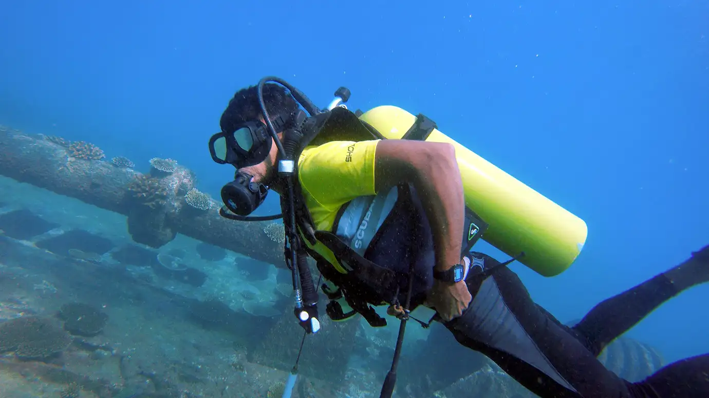 A scuba diver wearing a bright yellow short-sleeved shirt, black diving gear, and a yellow air tank swims through clear blue water past a submerged, coral-covered structure.