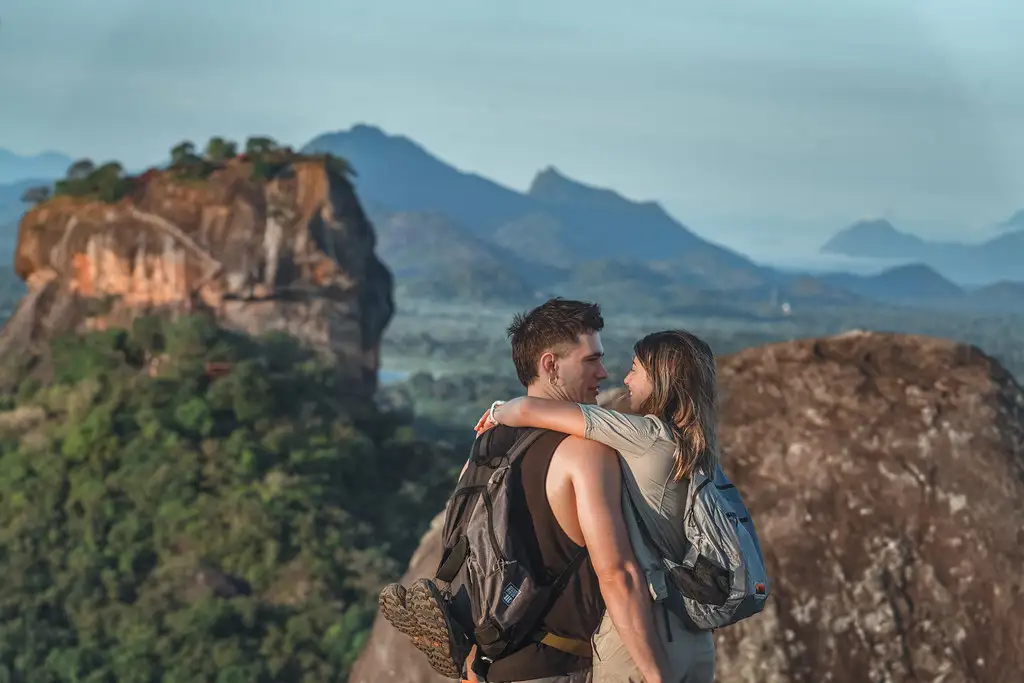 A couple wearing backpacks stands on a high rocky viewpoint, embracing and looking into each other's eyes. Behind them is a sprawling, lush green jungle landscape featuring the massive, flat-topped Sigiriya Rock Fortress on the left and distant blue mountains on the horizon.