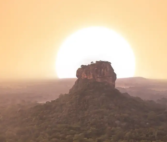 A massive, steep-sided rock formation stands in the center of a vast, dense green jungle. A giant, bright white sun sits perfectly behind the rock, creating a glowing halo effect against a vibrant, hazy orange and peach sky. A soft morning mist settles over the forest canopy below.