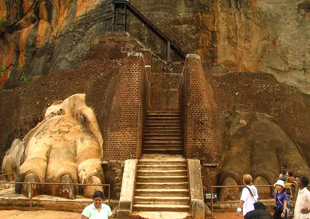 Tourists explore the ancient Lion's Paw entrance at Sigiriya Rock Fortress, a popular destination included in Sri Lanka Tour Packages. The image shows massive stone lion paws framing a central brick staircase leading up the sheer rock face.