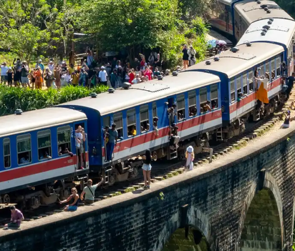 A blue and red passenger train travels across a curved stone arched bridge surrounded by lush green jungle. Large crowds of tourists stand on the grassy banks and along the tracks taking photos, while passengers lean out of the train's open doors and windows.