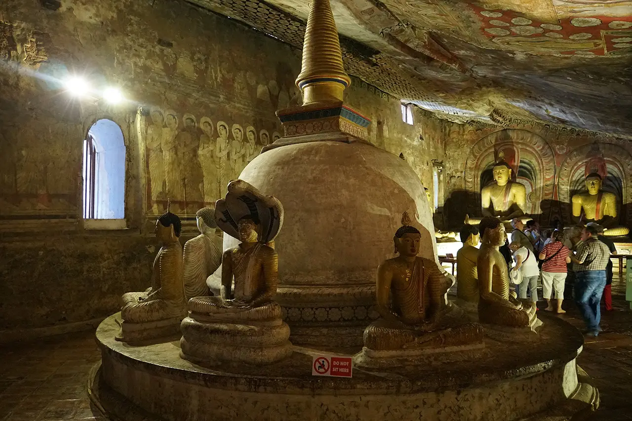 Visitors walking inside the sacred Dambulla Cave Temple, featuring a central stupa, multiple Buddha statues, and painted murals.