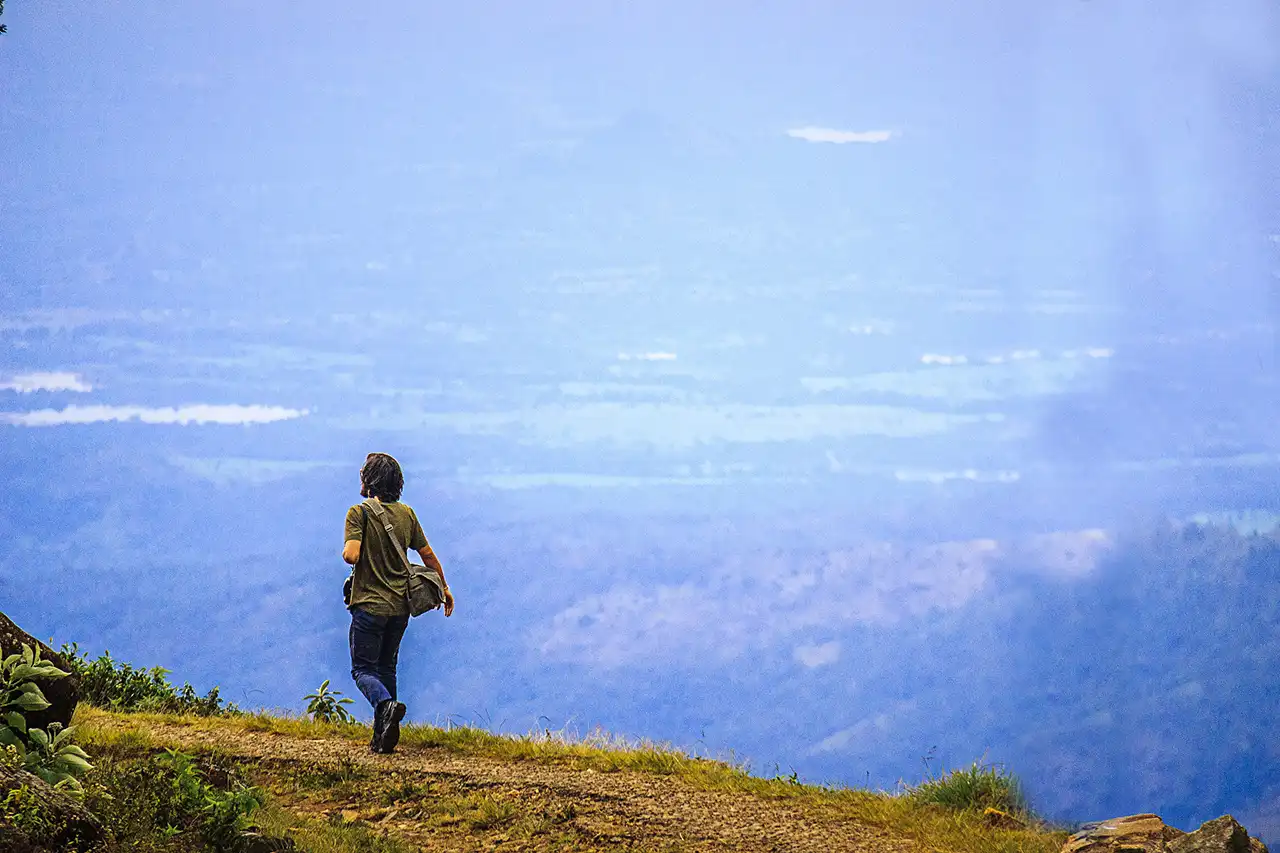 A hiker walking along a mountain ridge trail overlooking vast, misty valleys at Udaweriya in Sri Lanka.