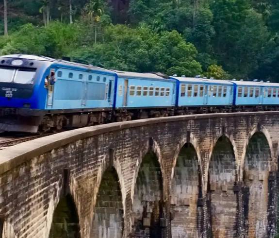 A blue train crossing the historic stone Nine Arch Bridge surrounded by lush green jungle in Sri Lanka.