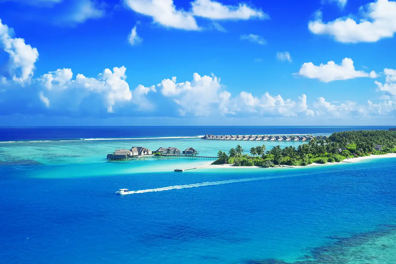 An aerial view of a tropical Maldivian island surrounded by vibrant turquoise lagoons, white sand beaches, overwater bungalows, and a small boat leaving a wake in the deep blue sea under a sky scattered with puffy white clouds.