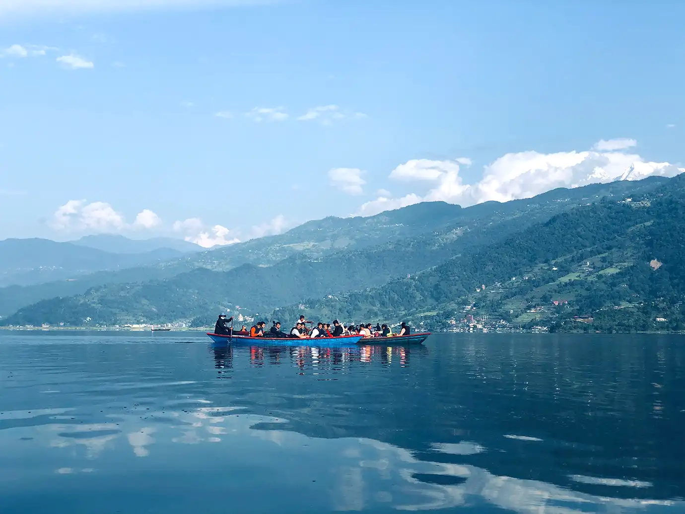 A wide landscape view of passengers riding in long wooden boats across a calm blue lake, framed by lush green hills, distant snow-capped mountains, and soft cloud reflections in the water.
