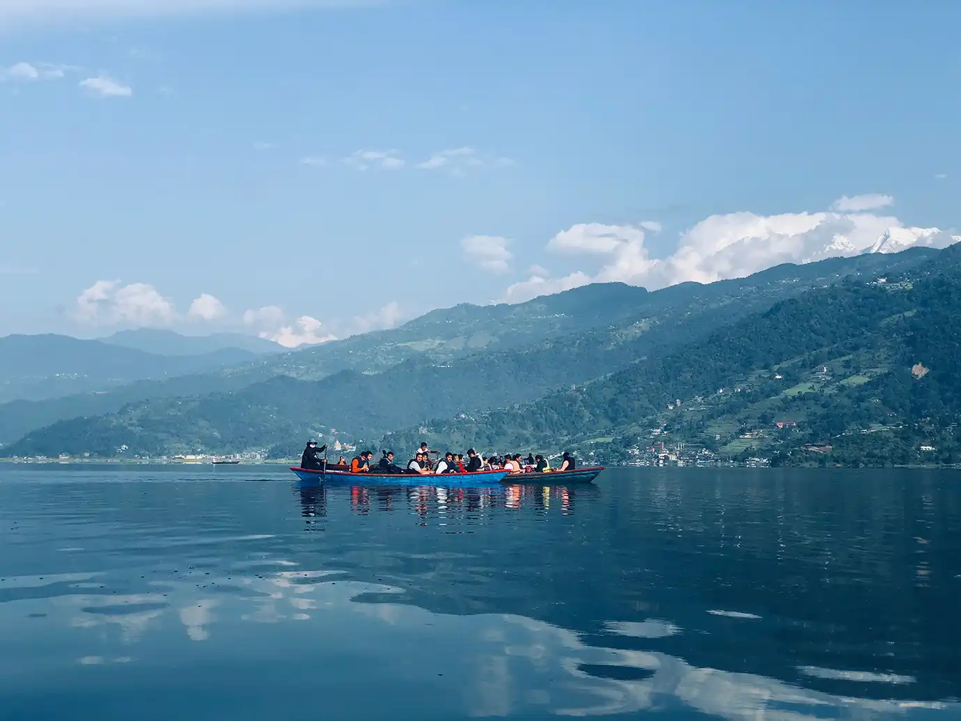 A wide landscape view of passengers riding in two long wooden boats across a calm blue lake, framed by lush green hills, distant snow-capped mountains, and soft cloud reflections in the water.