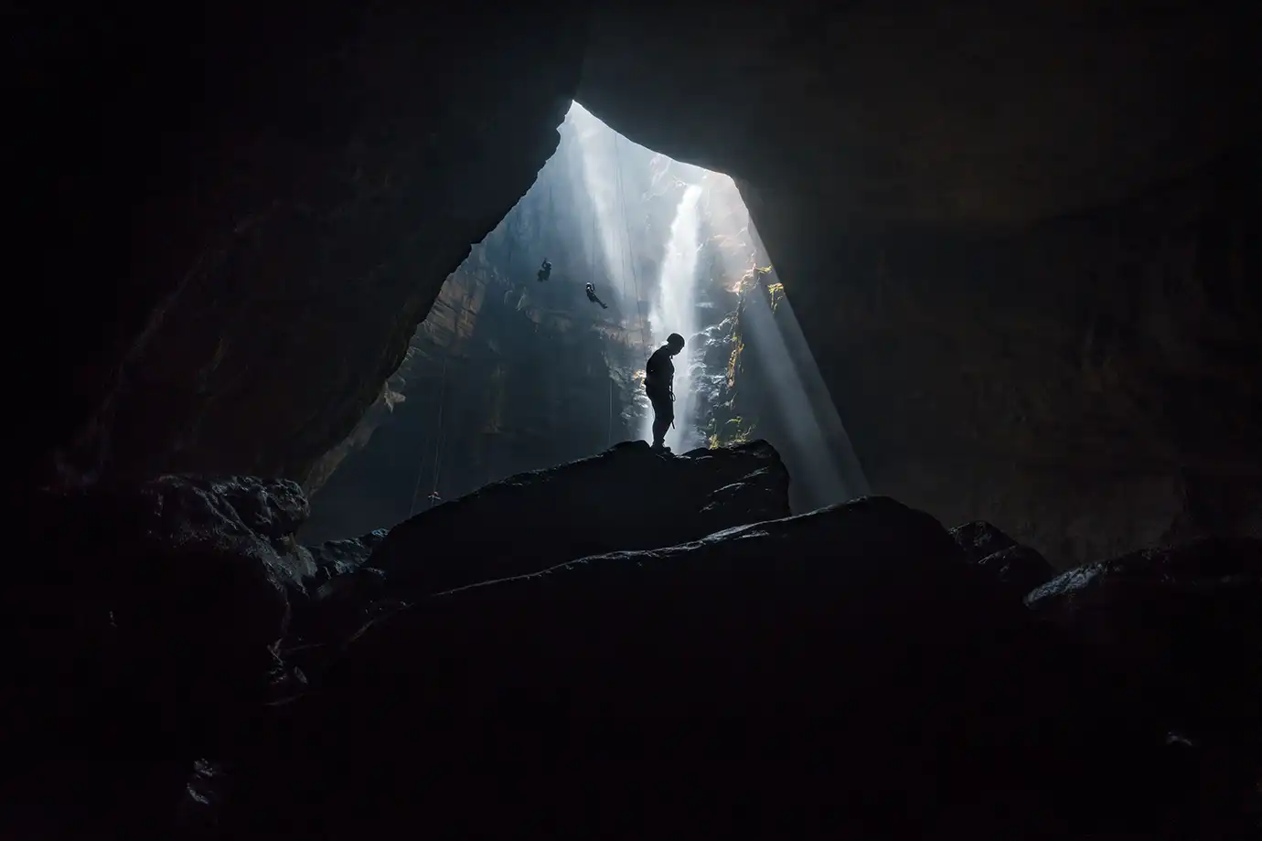 A dark silhouette of a person standing on a rocky outcrop inside a cavern, illuminated by a beam of sunlight streaming down from an opening above near a cascading underground waterfall.