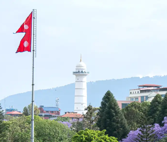 the iconic white Dharahara tower and the flying national flag of Nepal surrounded by lush greenery and blooming trees.