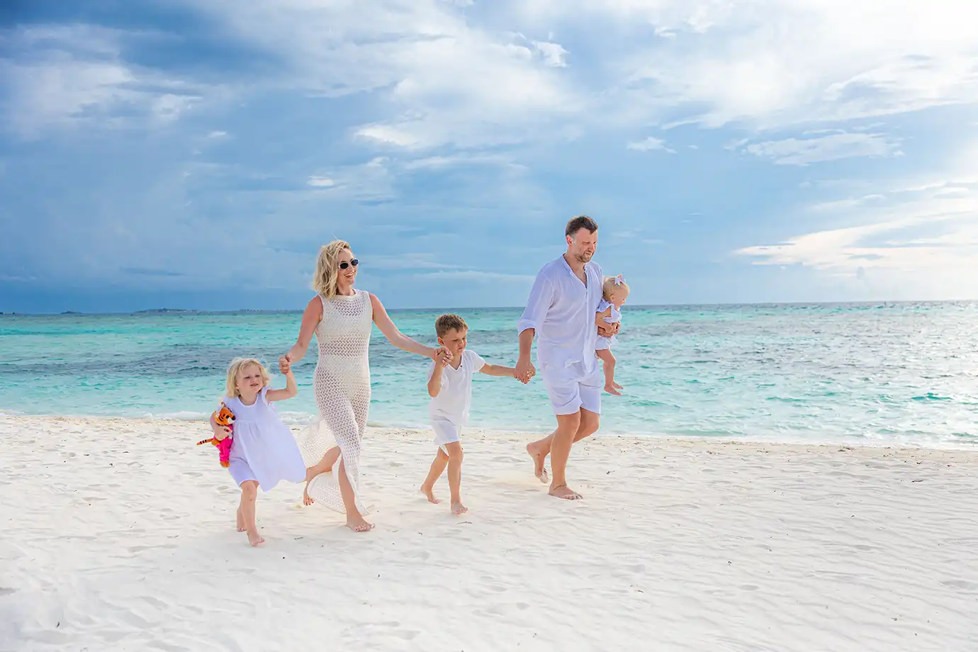 A family of five dressed in matching white outfits walks and plays together along a white sand beach beside clear turquoise ocean water under a cloudy blue sky.