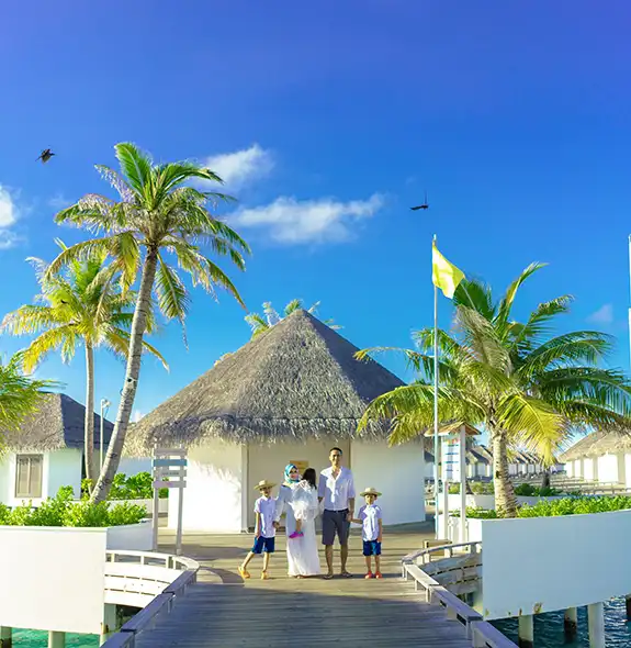 A family of five standing together on a wooden boardwalk lined with thatched-roof overwater villas and palm trees, under a bright blue sky with birds flying overhead in the Maldives.