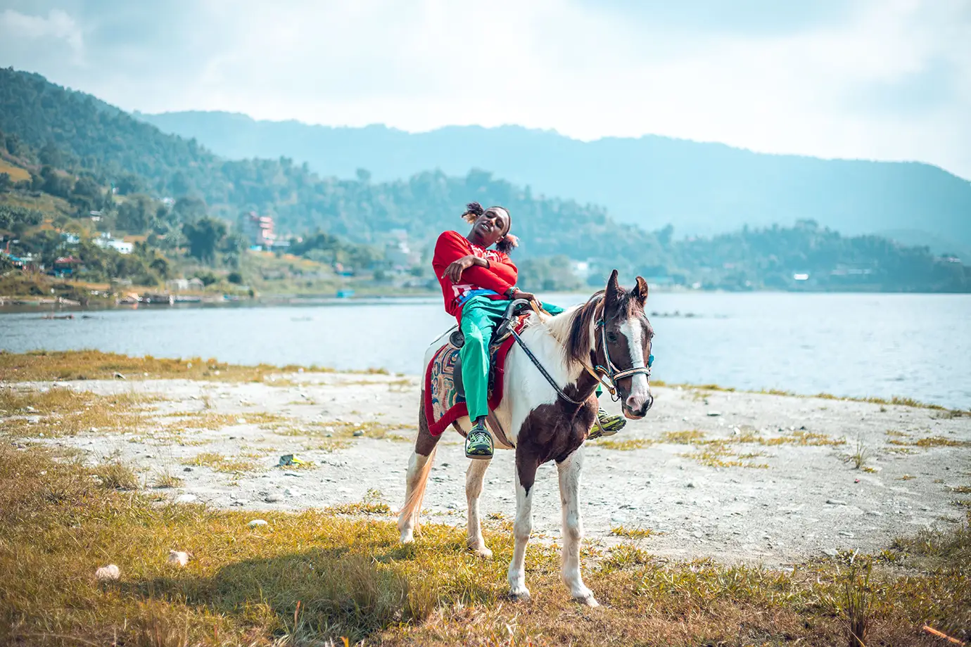 A young man wearing a red sweatshirt and green pants leans back relaxed on a brown and white horse by the grassy shore of Fewa Lake in Pokhara, with calm waters and misty green mountains in the background.