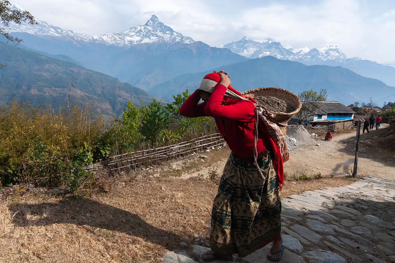 A woman dressed in a bright red sweater and patterned skirt carries a woven doko basket using a head strap along a stone village path, set against a backdrop of blue mountains and the snow-capped peak of Machhapuchhre in Nepal.