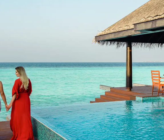 Two women in long flowing dresses, one white and one red, hold hands while walking along a wooden dock past a private infinity pool toward the turquoise ocean at a thatched-roof resort villa in the Maldives.