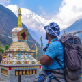 A trekker with a large backpack stands near a colorful stone stupa decorated with prayer wheels, gazing up at a cloud-swirled snow-capped peak in the Nepalese Himalayas where Nepal trekking permits are required.