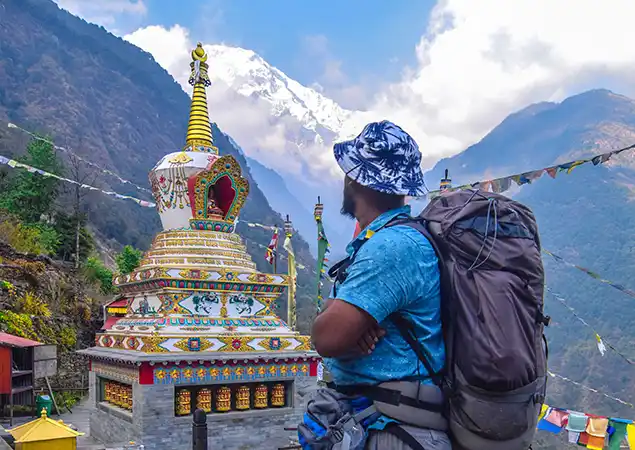 A trekker with a large backpack stands near a colorful stone stupa decorated with prayer wheels, gazing up at a cloud-swirled snow-capped peak in the Nepalese Himalayas where Nepal trekking permits are required.