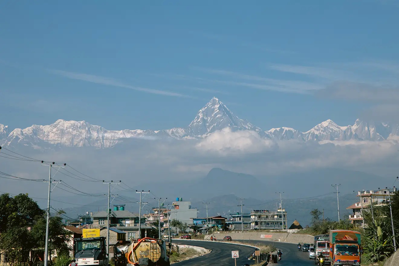 A wide paved highway in Nepal filled with trucks and utility lines, leading toward a distant city, with a thick layer of clouds hovering below the sharp, snow-covered peak of Machhapuchhre and the Annapurna mountain range under a clear blue sky.