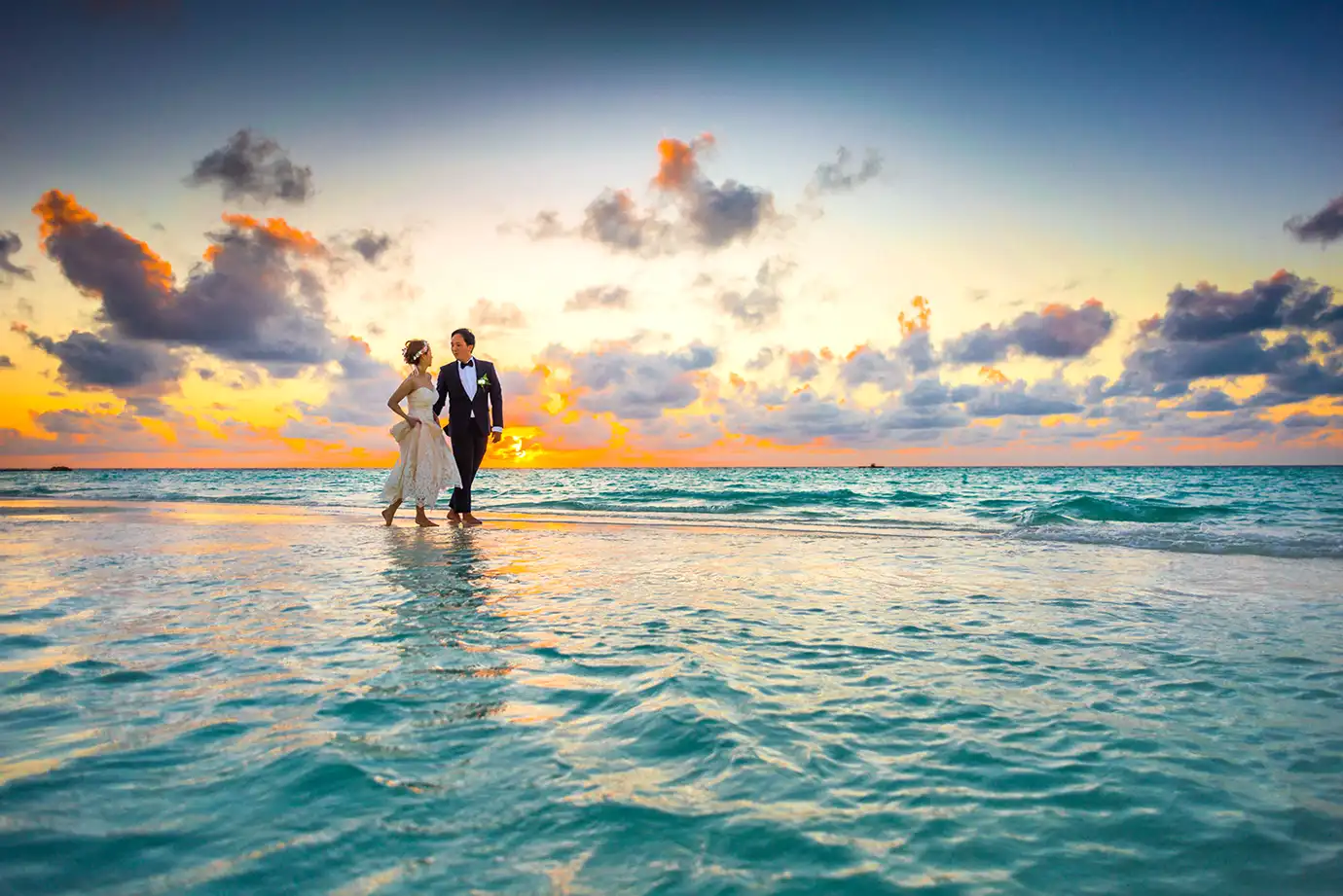 A bride in a white strapless wedding gown and a groom in a dark suit walk hand-in-hand along a shallow ocean shoreline at sunset, with golden and orange clouds illuminated across the sky.