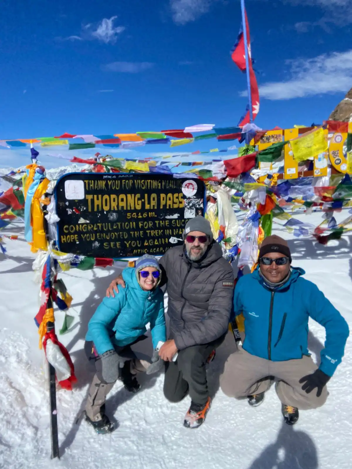 Best Time to Visit Nepal features smiling trekkers and their guide posing by the sign at Thorong La Pass at 5416 meters amidst snow and prayer flags.