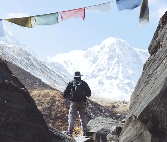 A lone trekker carrying a backpack stands on a rocky mountain path between two large dark stone formations, looking out toward a snow-capped Himalayan peak under colorful Buddhist prayer flags stretched across the sky.