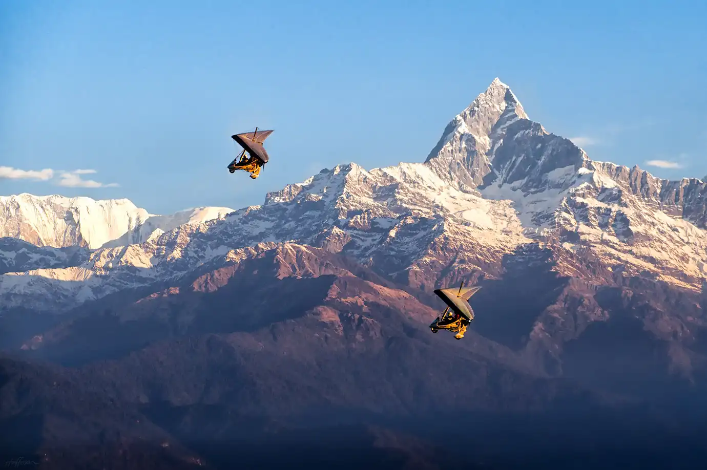 Two yellow ultralight trikes soar high in a clear blue sky, framed against the massive, snow-dusted peaks of Machhapuchhre and the surrounding Annapurna mountain range in Pokhara, Nepal.