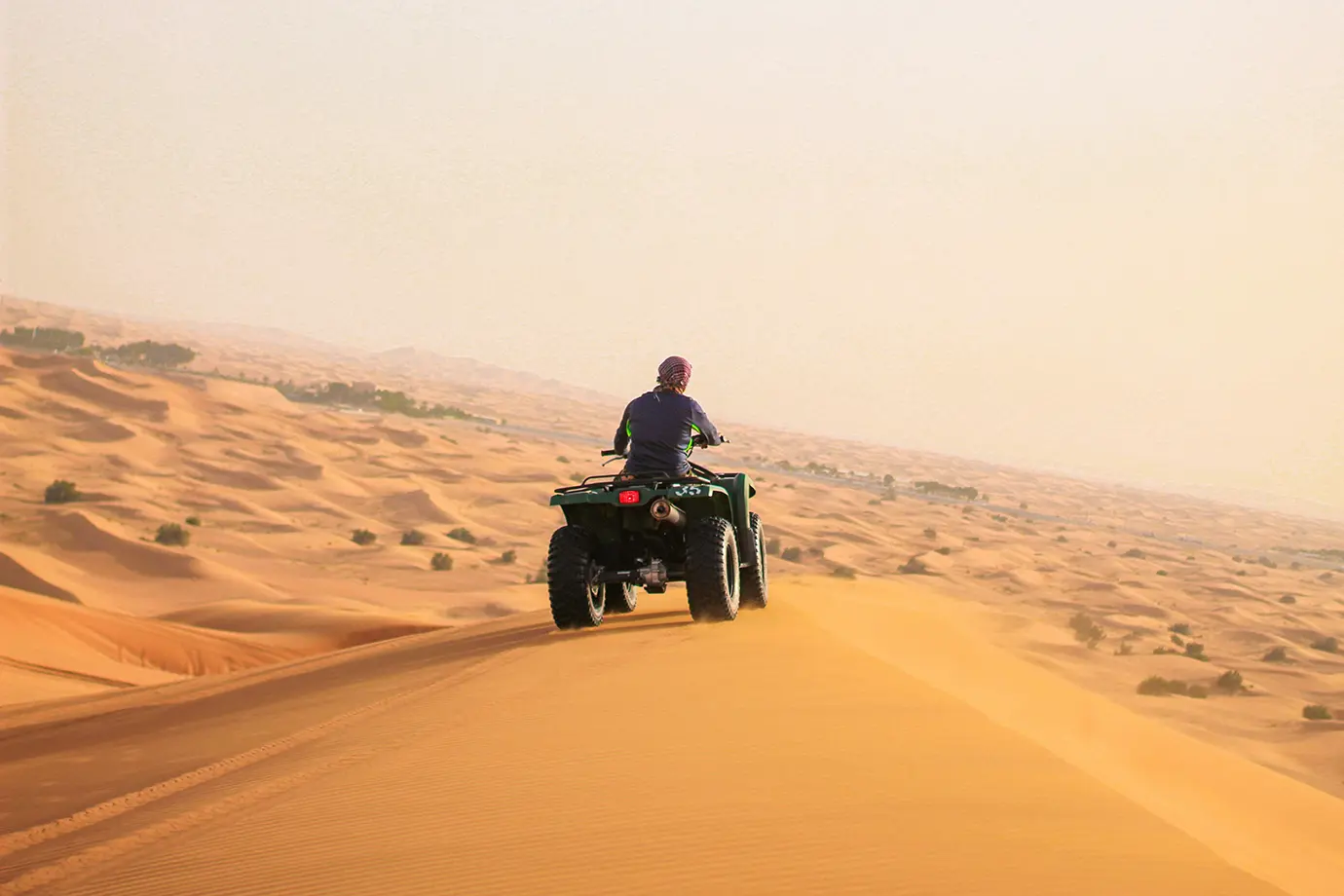 A person riding a green ATV quad bike along the high ridge of a golden sand dune during a Dubai Desert Safari.