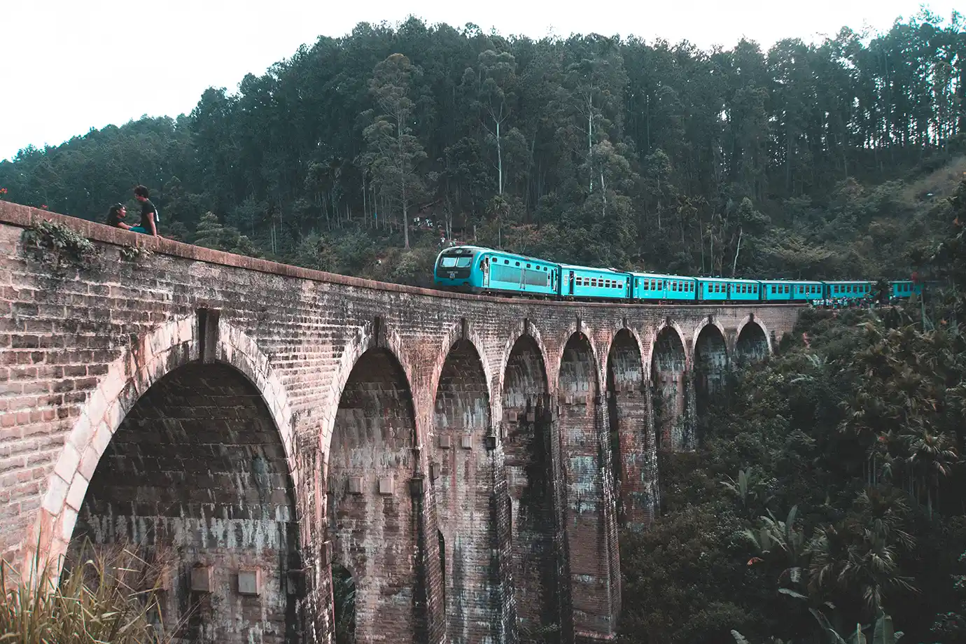 A low-angle photograph looking up at a bright blue passenger train crossing the stone arches of the Nine Arch Bridge surrounded by dense forest in Ella, Sri Lanka.