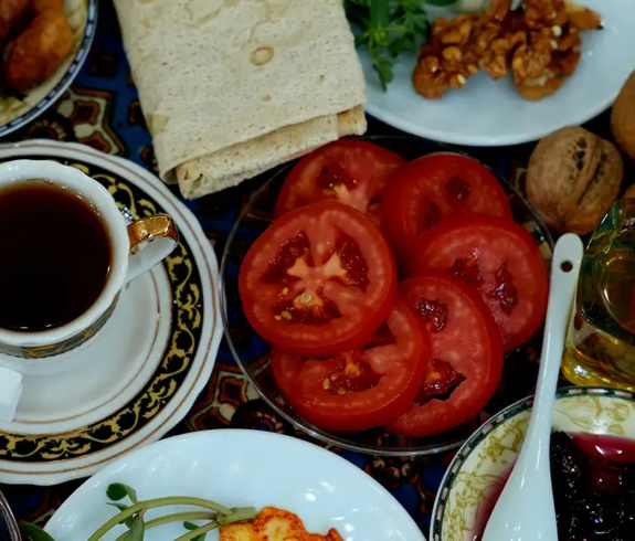A colorful flat-lay table spread featuring black tea, lavash flatbread, sliced tomatoes, walnuts, and local fruit preserves on an ornate tablecloth featured in a travel guide on Azerbaijani Food.