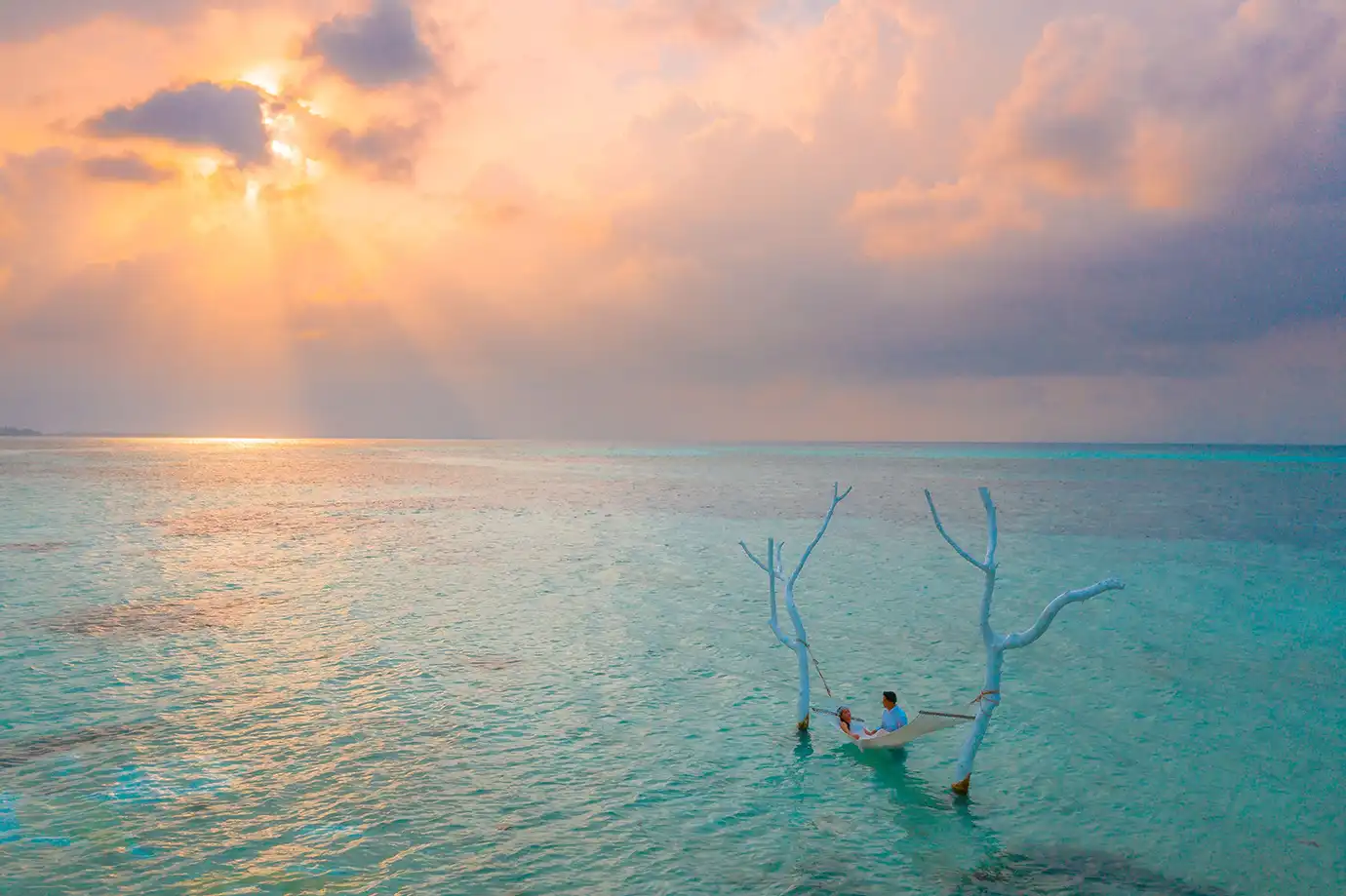 A couple lounging in a hammock suspended over calm sea waters between white wooden branches at sunset featured in an article asking Is a Maldives All-Inclusive Luxury Resort Holiday Worth It?