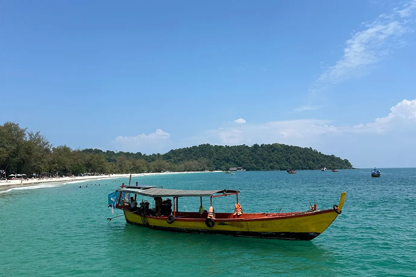 A colorful yellow and red wooden longtail boat floating on clear turquoise ocean water near a white sandy beach in Cambodia featured in a travel guide on the Best Time to Visit Cambodia.