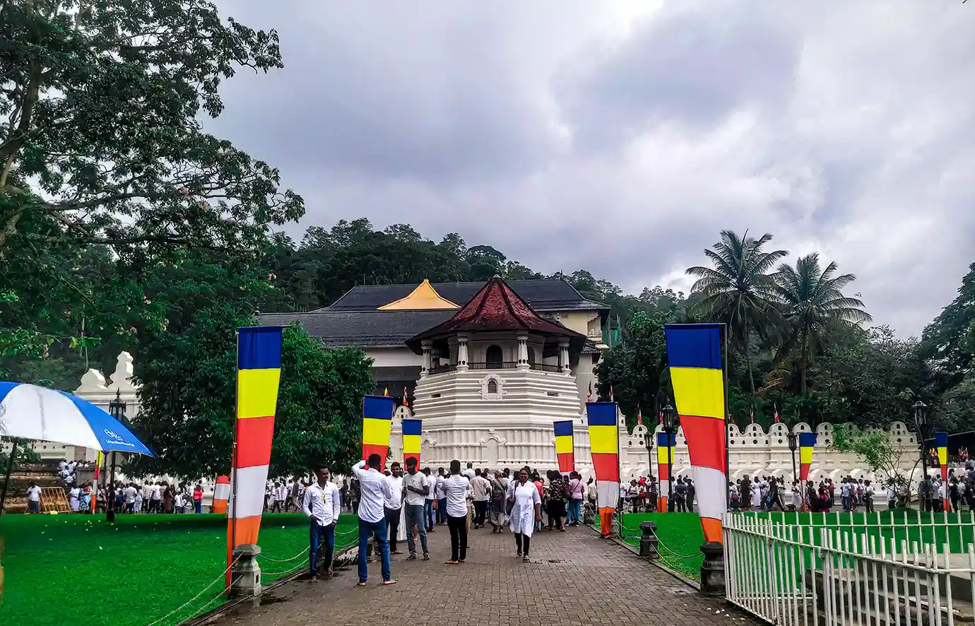 A bustling crowd of visitors walk along a paved path lined with colorful Buddhist flags toward the iconic white Octagon at the Temple of the Tooth in Kandy.