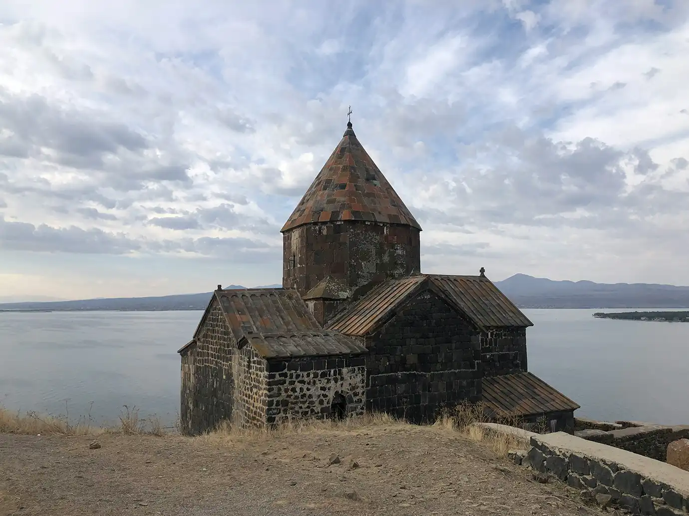 Medieval black stone church of Sevanavank Monastery set against Lake Sevan under a cloudy sky featured in a travel guide on When Is the Best Time to Visit Armenia.