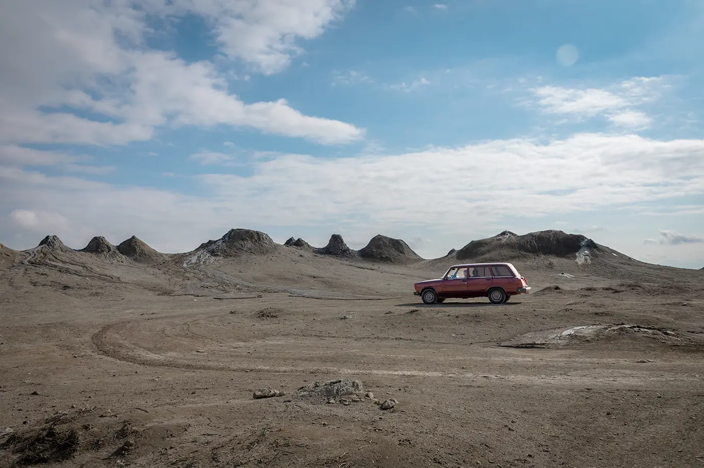 A reddish-pink vintage Soviet Lada car parked on dirt ground beside mud volcano cones in Gobustan featured in a travel guide on How Much Does a Trip to Azerbaijan Cost?