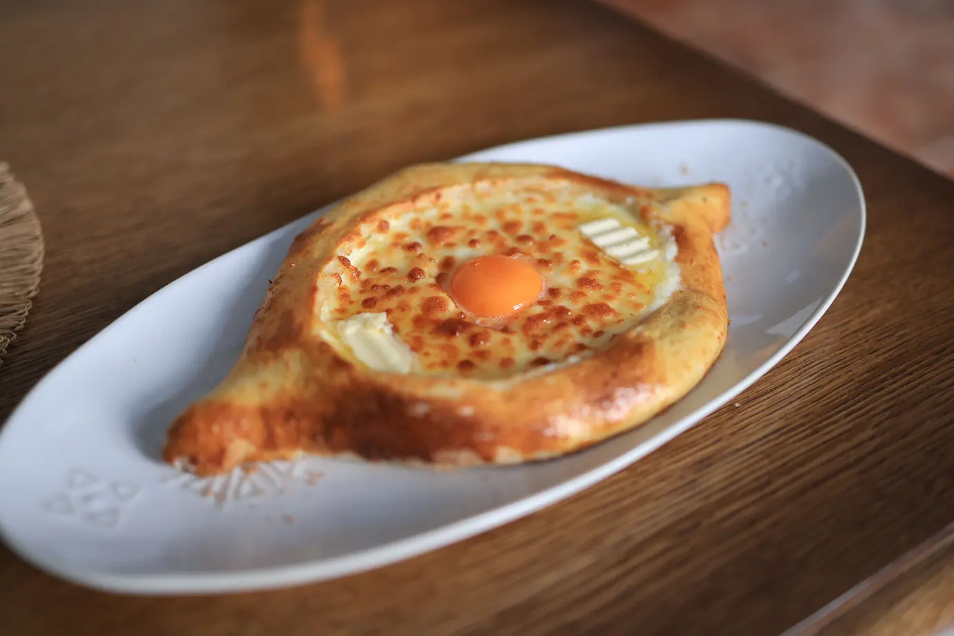 A golden boat-shaped Adjaruli Khachapuri filled with melted cheese, a pat of butter, and a raw egg yolk on a white oval plate, highlighting staple recommendations for a Georgian Food Guide.