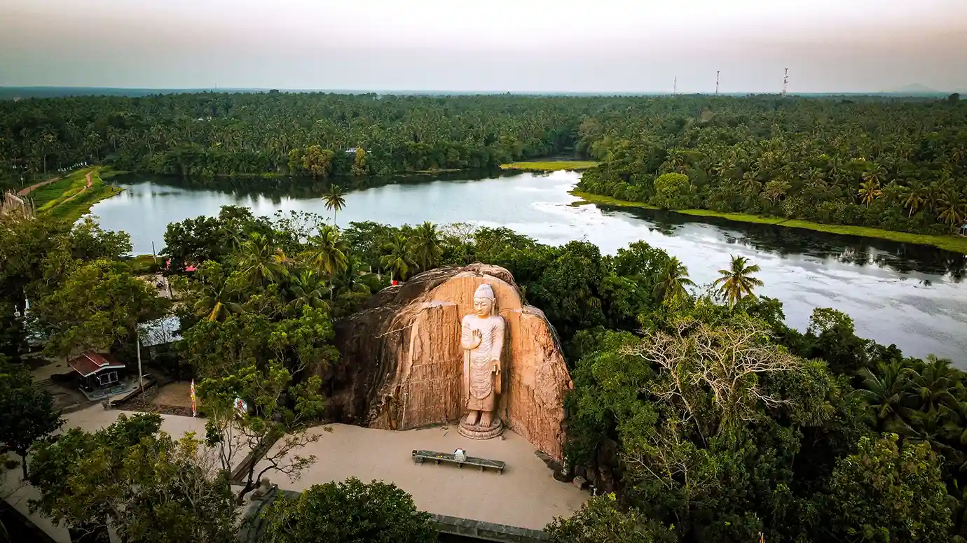 High-angle aerial shot of the standing Avukana Buddha statue surrounded by dense tropical forest and a large reservoir.